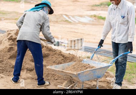 Worker pours sand from a large pile of sand onto a construction wheelbarrow in building site. Stock Photo