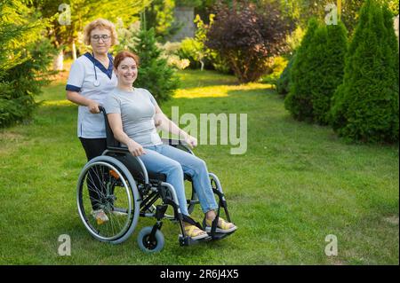 An elderly female nurse walks with a middle-aged woman in a wheelchair in the park.  Stock Photo