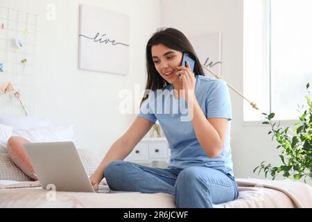 Female freelancer with laptop talking by mobile phone in bedroom Stock Photo
