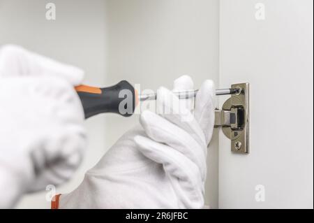 A man in gloves using a screwdriver assembles new kitchen furniture at his home. a person in white gloves holding a screwdriver Stock Photo
