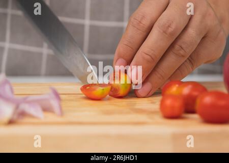 Female hand using a knife to slice fresh tomatoes on a cutting board. Close-up. Woman cutting cherry tomatoes at home. Preparing homemade food Stock Photo