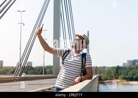 A male tourist with a backpack in sunglasses and a straw hat stands on a bridge over the river and takes a selfie using his phone. Stock Photo
