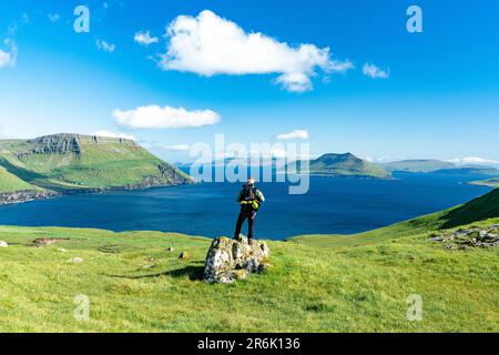 Hiker standing on top of rock contemplating the fjord in summer, Nordradalur, Streymoy Island, Faroe Islands, Denmark, Europe Stock Photo