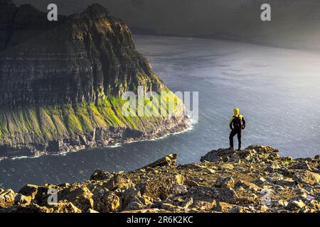 One man admiring the majestic cliffs along a fjord standing on top of mountain during a hike, Vidoy Island, Faroe Islands, Denmark, Europe Stock Photo