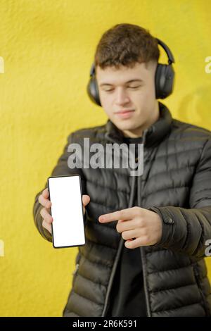 Caucasian boy with headphones out of focus shows the blank screen of his mobile phone isolated on a yellow background. Stock Photo