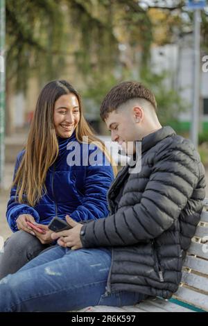 Couple of girl and boy sitting on the bench in a public park look at the mobile phone. Stock Photo