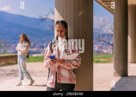 Brunette school girl using calculator for her homework while leaning on a pillar at the school yard. Her friend is having phone call in the back Stock Photo