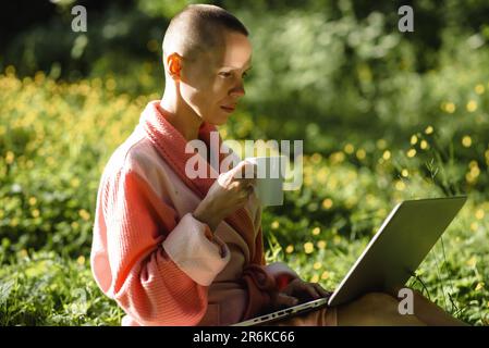 A bald woman sits with a laptop on the green grass and drinks coffee. Freelancer, work in nature. Stock Photo