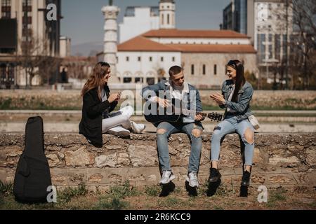 Handsome boy is playing guitar while sitting with his two girlfriends near the river Stock Photo