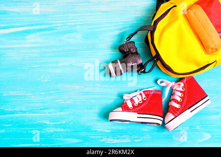 Tiny red canvas shoes, colorful backpack, and toy camera replica on a blue wooden background with copy space. Back-to-school deals and promotions conc Stock Photo