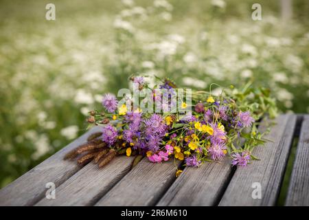 A wreath of wildflowers on a wooden garden table against the backdrop of a green flowering meadow Stock Photo
