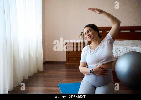 Delightful gorgeous pregnant woman holds her hand on her big belly, stretch her arm righ, practices prenatal stretching Stock Photo