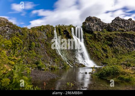 A pristine waterfall cascades down the face of a majestic mountain, the sunny sky reflecting off the clear waters Stock Photo