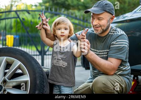 Cute toddler boy helping his father to change car wheels at their backyard. Father teaching his little son to use tools. Active parent of a small chil Stock Photo