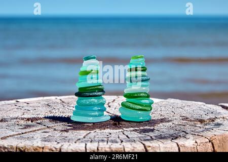 Two balanced pyramids of wet, sea-polished fragments of a blue and green glass bottle on a weathered wooden trunk cut against the sea Stock Photo
