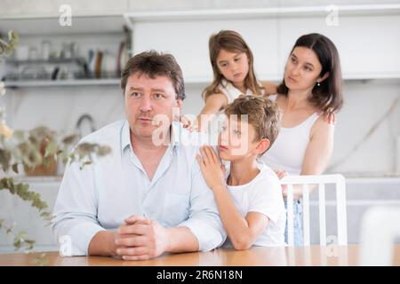 Offended father sitting at the kitchen table being hugged by son Stock Photo