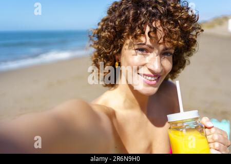 Cheerful woman enjoying juice and taking self portrait on beach Stock Photo