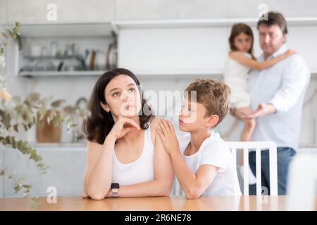 Offended mother sitting at the kitchen table being hugged by son Stock Photo