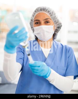 Female nurse wearing a protective mask fills a syringe with saline solution in the treatment room Stock Photo