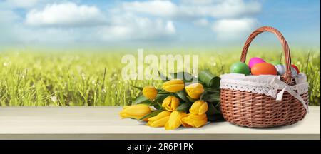 Wicker basket with festively decorated Easter eggs and tulips on wooden table on green meadow, space for text. Banner design Stock Photo