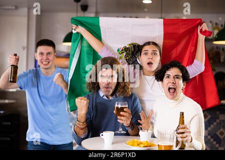 Company of emotional young adult sports fans supporting Mexican team with state flag while resting in pub with beer Stock Photo