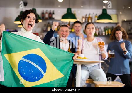 Company of emotional young adult fans supporting sports team of Brazil with state flag while resting with beer in bar Stock Photo