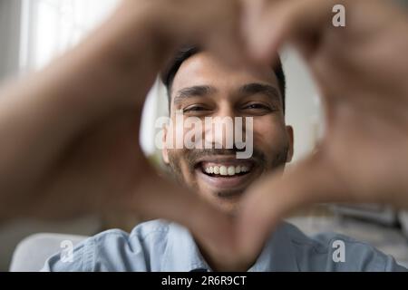 Cheerful young Indian man smiling at camera through hand heart Stock Photo