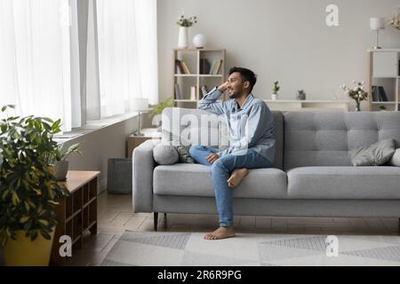 Positive dreamy relaxed young Indian guy sitting on comfortable couch Stock Photo