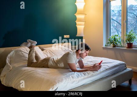 Young man lying on bed, using mobile phone, reads news, chatting, dating in social media Stock Photo