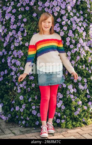 Outdoor portrait of pretty little girl wearing rainbow pullover and denim skirt, posing next to Morning Glory flowers wall Stock Photo