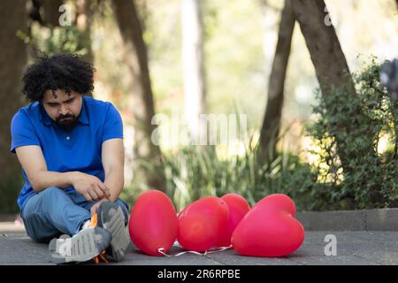 A young man in casual attire kneels on the pavement of a busy city street, surrounded by a colorful display of vibrant red balloons Stock Photo