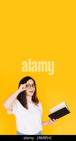 Portrait of a happy young woman wearing glasses and white T-shirt, immersed in reading a book with an open mouth, isolated on a yellow background Stock Photo