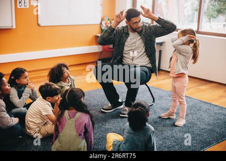 Male educator in a primary school class. Teacher shows a little girl how to do heads and shoulders. Man leading children on an educational activity in Stock Photo