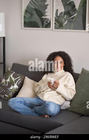 Young happy latin woman drinking tea or coffee relaxing at home on couch. Stock Photo