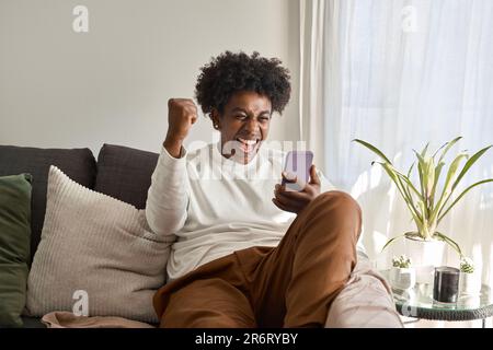 Excited African American guy sitting on couch holding phone celebrating win. Stock Photo