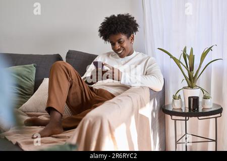 Happy gen z African American teen sitting on couch using mobile phone tech. Stock Photo