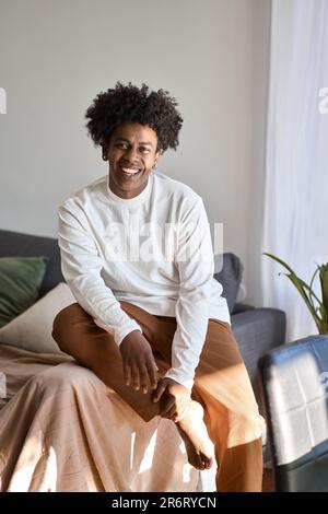Young smiling happy gen z African American guy sitting on couch at home. Stock Photo