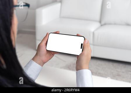 Young woman is sitting on a white couch, holding and watching smartphone in horizontal position. Isolated screen in white for mockup, app presentation Stock Photo