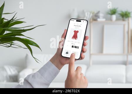 Woman shopping online with smart phone. Buying a red flow dress on ecommerce web page. Living room interior in background Stock Photo