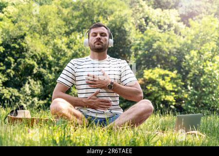 Man wearing headphones doing yoga breathing exercise in half lotus position in the park, listening music for meditation. Stock Photo