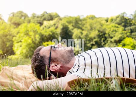 A man in sunglasses is resting in the park, lying on his back on the green grass and looking at the sky. Stock Photo