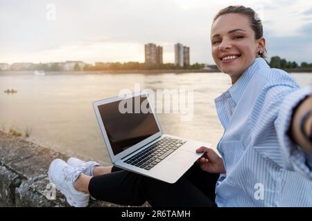 Young urban woman working out of the office sitting on waterfront meeting sunset and taking a selfie. Stock Photo