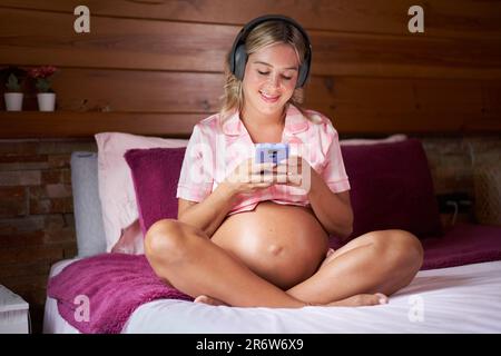 Smiling pregnant woman sitting on bed at home using cell phone. Cheerful Mom connected online. Stock Photo