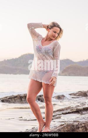 Blonde woman standing on beach in white crochet bathing suit cover in Nicaragua on the pacific ocean near San Juan del Sur at sunset in water Stock Photo