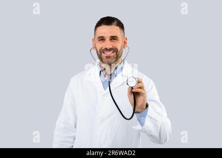 Friendly middle aged man doctor wearing white medical coat posing on light background, using stethoscope and smiling Stock Photo
