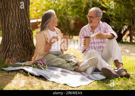 Happy european mature couple with cup of coffee takeaway, sandwich sit on plaid, enjoy date Stock Photo
