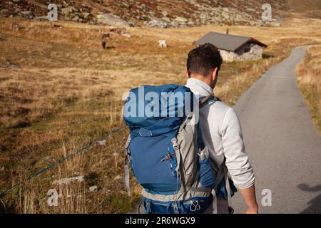 A young man with a backpack stands on a mountain road in Switzerland, gazing at the breathtaking view behind him Stock Photo