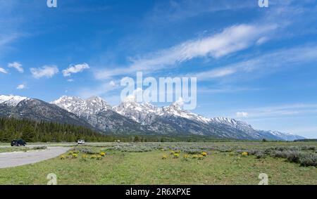 See the long road that runs below Grand Teton National Park in Wyoming, USA. Mountains are snow and high with cloud drifts above a blue sky. Stock Photo