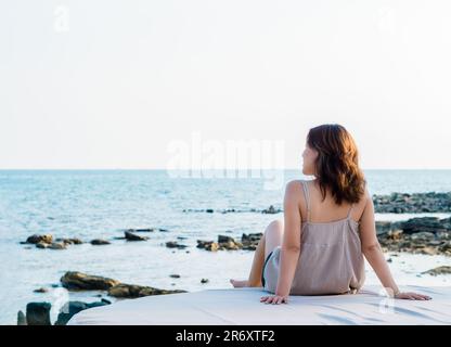 Back of Asian woman in casual dress sit in front of the beach in summer vibes. Lonely female relaxing smile at the sea and looking far away, seascape Stock Photo
