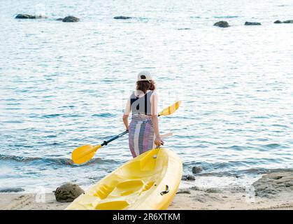 Asian woman in black tank top and cap dragging yellow kayak and holding paddle on the sand to the sea. Happy female having fun activity at the beach f Stock Photo